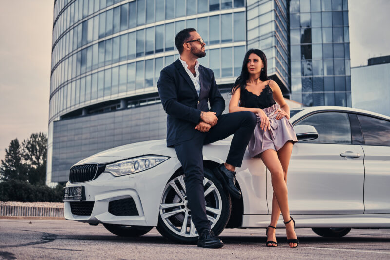 Well-dressed attractive couple leaning on a luxury car outdoors against a skyscraper.